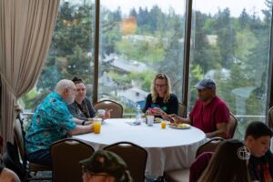 Four people are sitting at a round table enjoying a VIP brunch by a large window with a scenic view on this lovely Sunday.