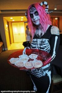 A person with pink hair and skeleton makeup holds a red tray with individually wrapped treats, ready to welcome platinum ticket holders at the exclusive Celebrity Meet and Greet.