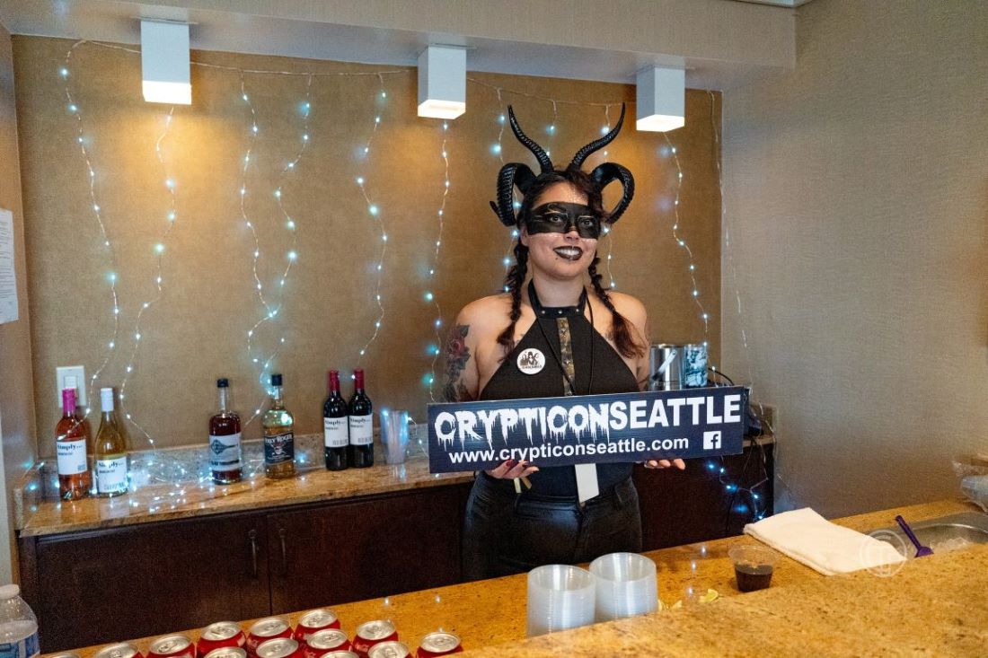 Person in a demon costume holding a "Crypticon Seattle" sign behind a counter, with drinks and fairy lights in the background. Platinum ticket holders can enjoy exclusive perks and perhaps even a surprise Celebrity Meet and Greet during the event.