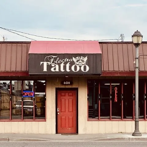 The "Electric Tattoo" shop presents a striking front view with its bold red door. Illuminated open signs adorn the windows, creating a welcoming ambiance. A contrasting white logo features prominently on the black awning, uniquely identifying the business.