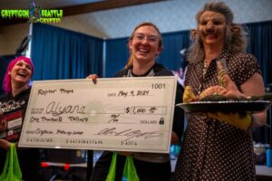 Three people on stage at Crypticon Seattle, one holding a giant $1,000 check, another in costume holding a turtle—part of the thrilling Makeup Contest.
