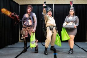 Three people in elaborate costumes, celebrating on stage at Crypticon Seattle with green gift bags in hand.