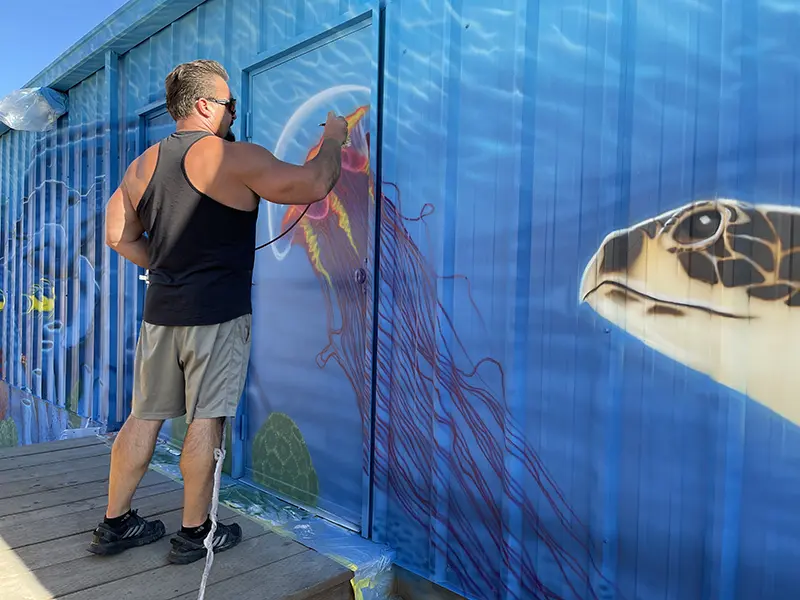A man is attending a Dutch airbrush boot camp class where he is involved in the task of painting a wall.