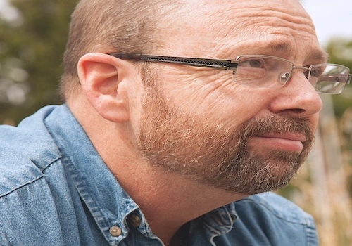 A bearded man with glasses gazes thoughtfully into the distance, perhaps pondering media mysteries in an outdoor setting.