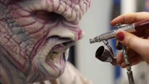 A person airbrushing intricate alien makeup on another person's face.