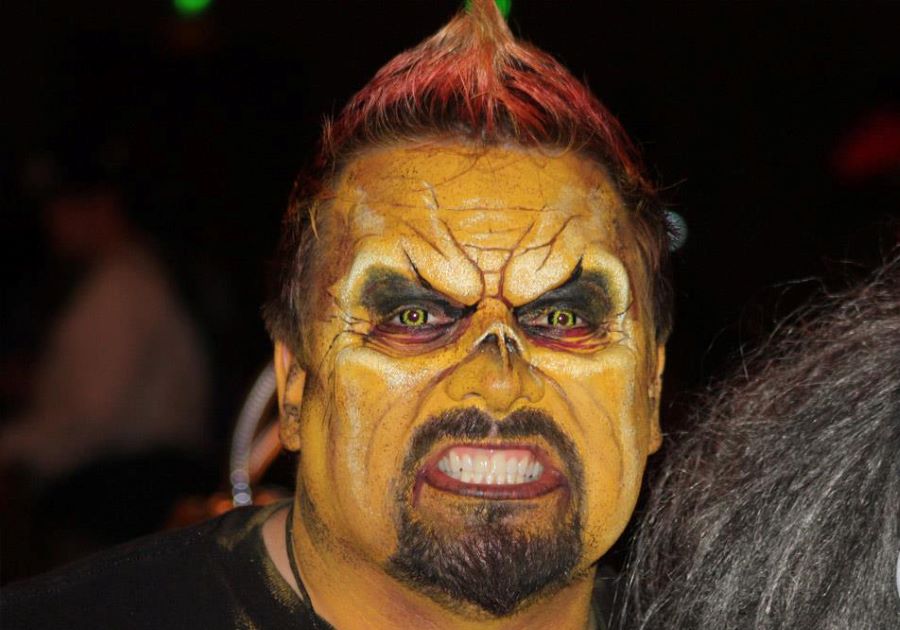 Person with detailed, monster-like SFX makeup, yellow skin, dark eyes, and a fierce expression against a dark background.