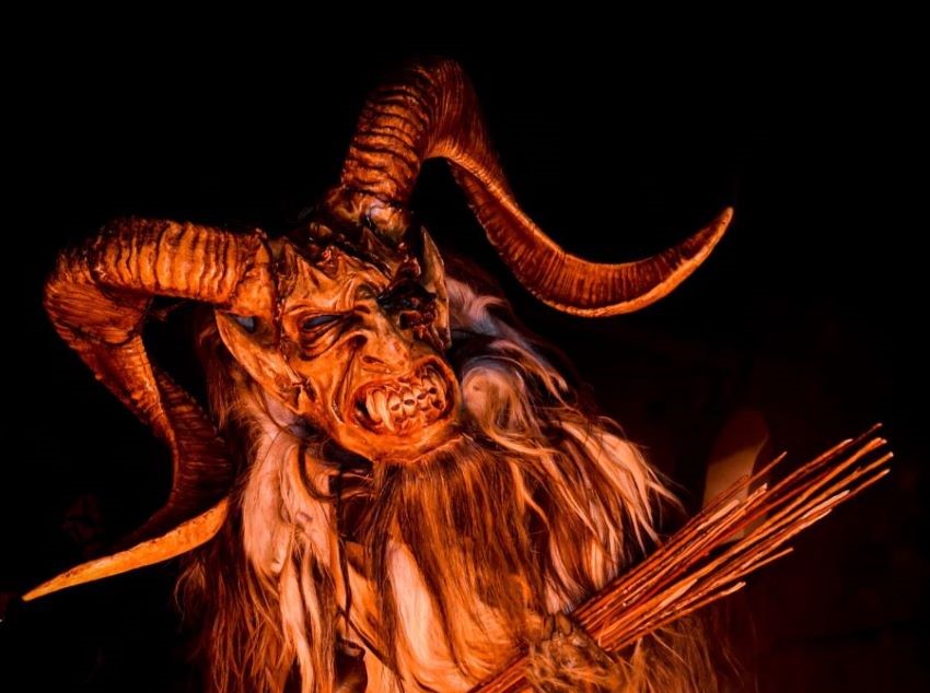 A Bremerton Krampus in a horned costume stands behind flames, holding a bundle of sticks.
