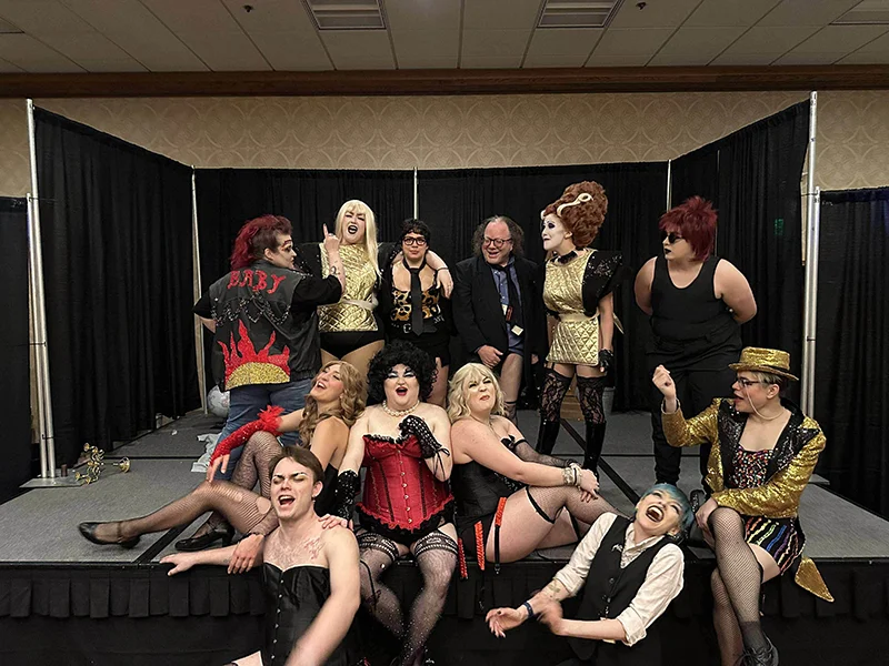 A group in costume poses on stage, some sitting, some standing, with playful and dramatic expressions.