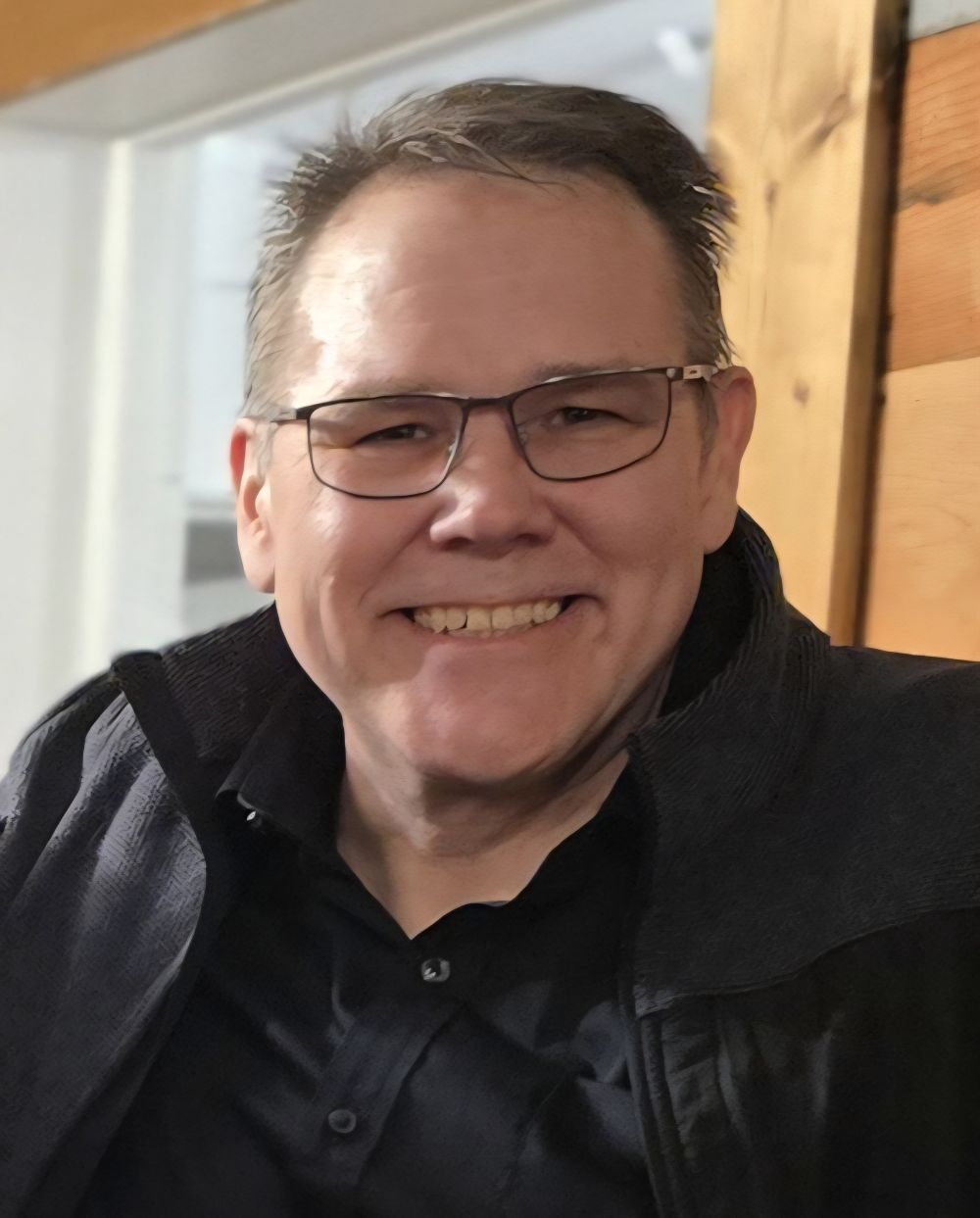 Smiling man with glasses and short hair, seated indoors by a wooden wall at the Costume Workshop.