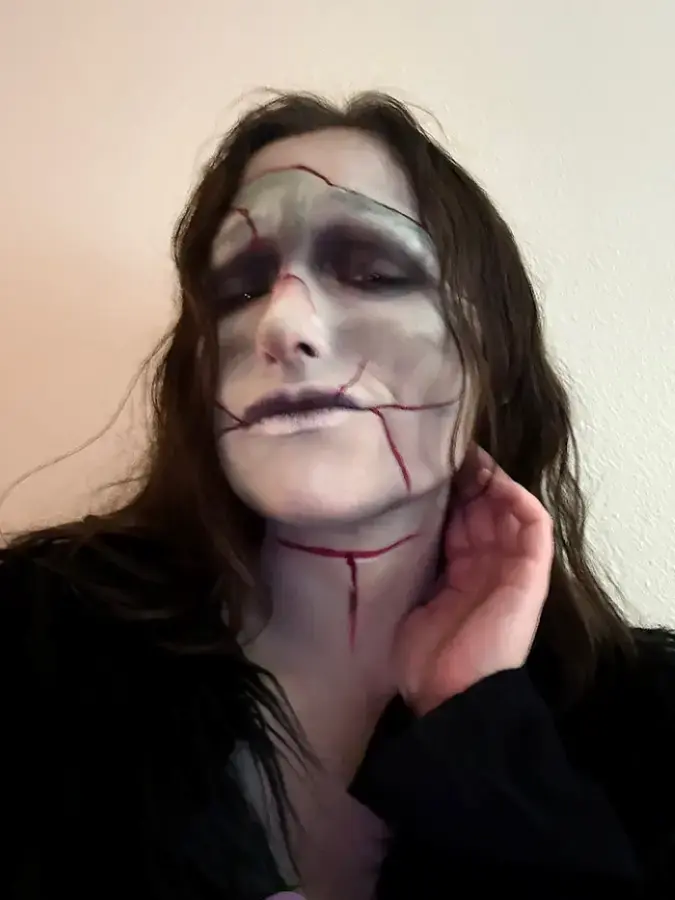 Person with long hair wearing cracked doll face makeup and dark clothing, posing with hand near neck.