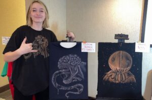 Zayda smiling and posing with two surreal creature art Bleach T-Shirts and price signs.