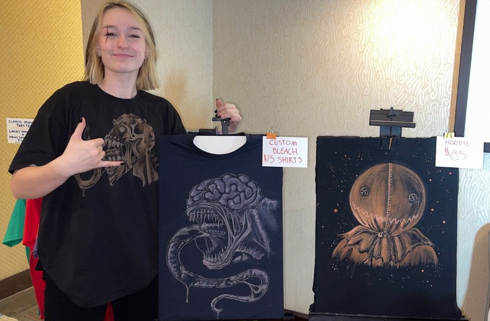 Smiling Zayda poses with two black bleach t-shirts featuring hand-painted monster designs and price signs.