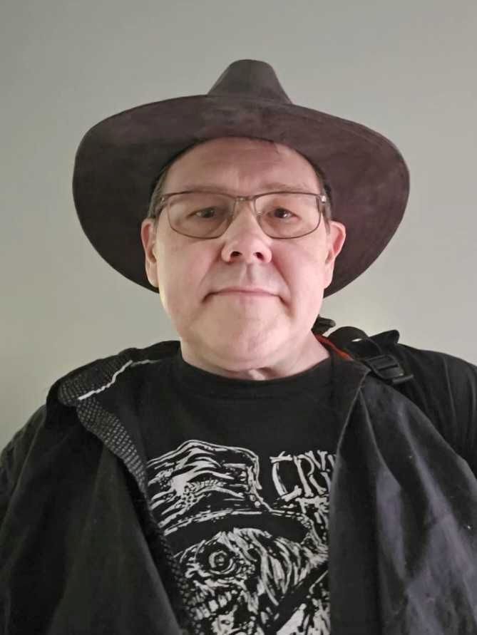 Robert Buchta wears glasses and a wide-brimmed hat, dressed in a black graphic t-shirt and jacket, facing forward in this demo.