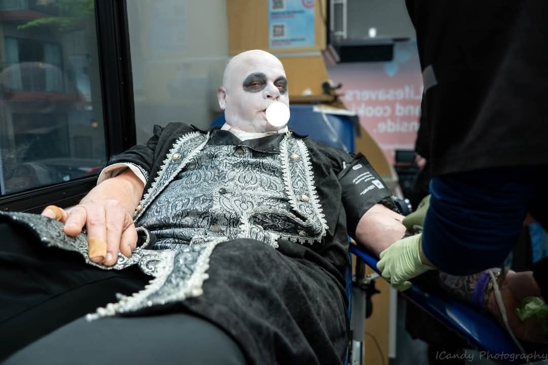 Person in ornate costume with white face paint donates blood at a Blood Drive, blowing bubble gum, attended by a gloved worker.