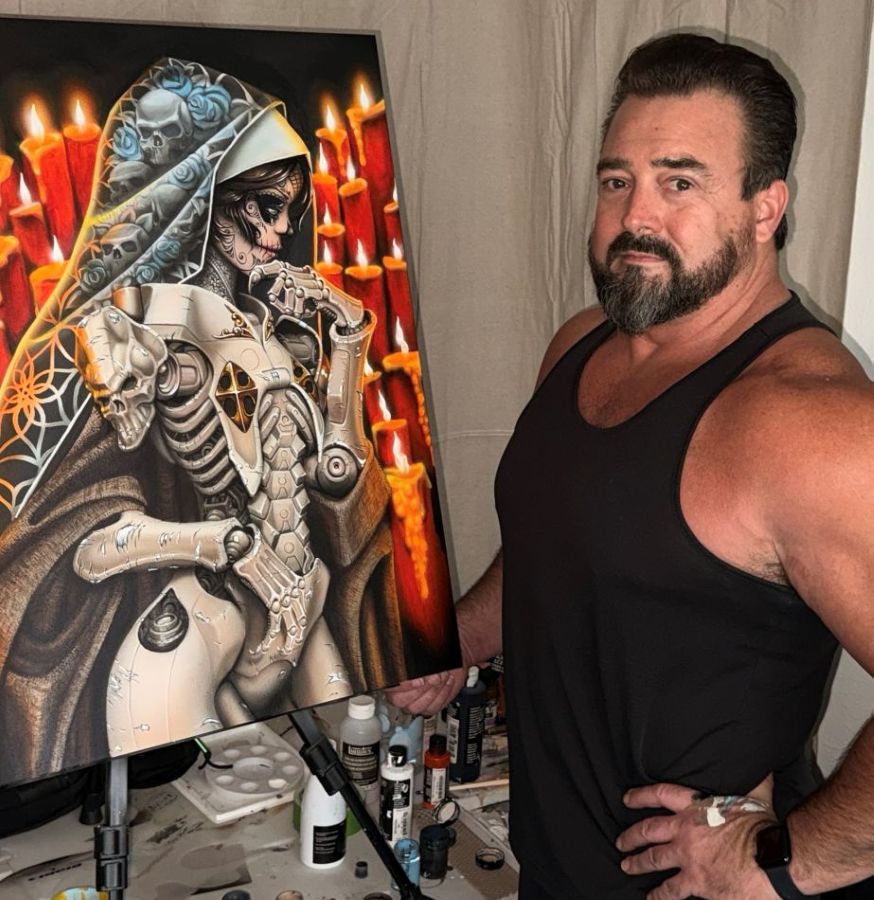 A man in a black tank top stands by a Dutches Demo painting of a robotic, skeletal nun with candles in the background.