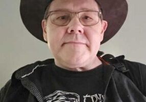 Robert Buchta wears glasses and a wide-brimmed hat, dressed in a black graphic t-shirt and jacket, facing forward in this demo.