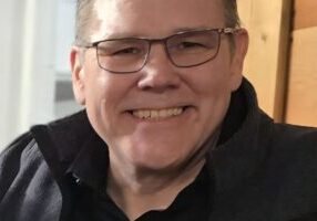 Smiling man with glasses and short hair, seated indoors by a wooden wall at the Costume Workshop.
