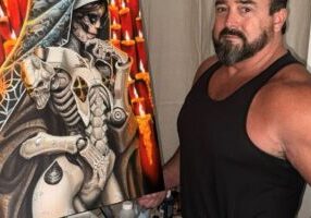 A man in a black tank top stands by a Dutches Demo painting of a robotic, skeletal nun with candles in the background.