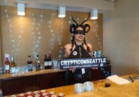 Person in a demon costume holding a "Crypticon Seattle" sign behind a counter, with drinks and fairy lights in the background. Platinum ticket holders can enjoy exclusive perks and perhaps even a surprise Celebrity Meet and Greet during the event.