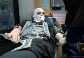 Person in ornate costume with white face paint donates blood at a Blood Drive, blowing bubble gum, attended by a gloved worker.
