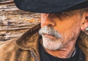 cowboyhowell Older man with a grey mustache in a black cowboy hat and brown jacket, like C. Thomas Howell in an interview, stands by a wooden wall.