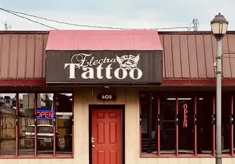 The "Electric Tattoo" shop presents a striking front view with its bold red door. Illuminated open signs adorn the windows, creating a welcoming ambiance. A contrasting white logo features prominently on the black awning, uniquely identifying the business.