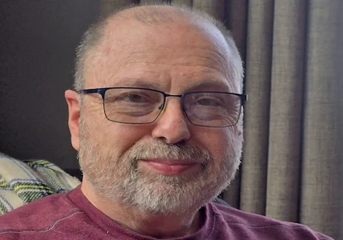 Horrorwriter Mark Allen, an older man with glasses and a beard, smiles indoors near a window with curtains in his maroon shirt.