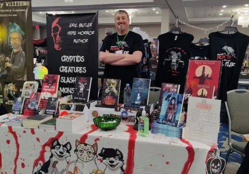 Eric Butler sits at a convention table with his books, posters, themed t-shirts, and a red splattered tablecloth.