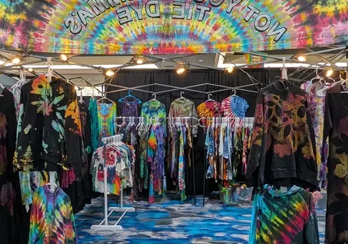 A vibrant Tie Dye booth at the market, displaying colorful shirts, jackets, and dresses on racks.