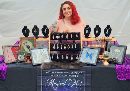 A woman with red hair smiles behind a Magical Me Gems display of gemstone jewelry and oddities at her booth.