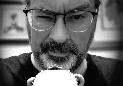 Man with glasses and a beard winking while holding a cup close to his face in black-and-white.