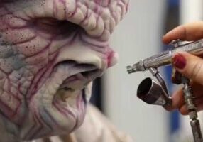 A person applies airbrush makeup to a face with detailed alien prosthetics.