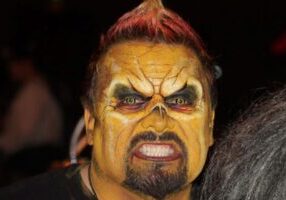 Person with detailed, monster-like SFX makeup, yellow skin, dark eyes, and a fierce expression against a dark background.