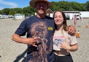 A man in a straw hat and Zayda smile and pose for a photo outside on a sunny day.